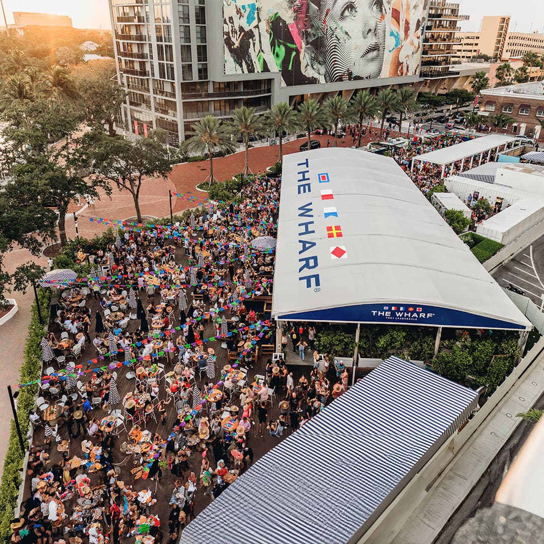Aerial view of The Wharf Fort Lauderdale venue at sunset with crowds under colorful bunting and the signature white tent