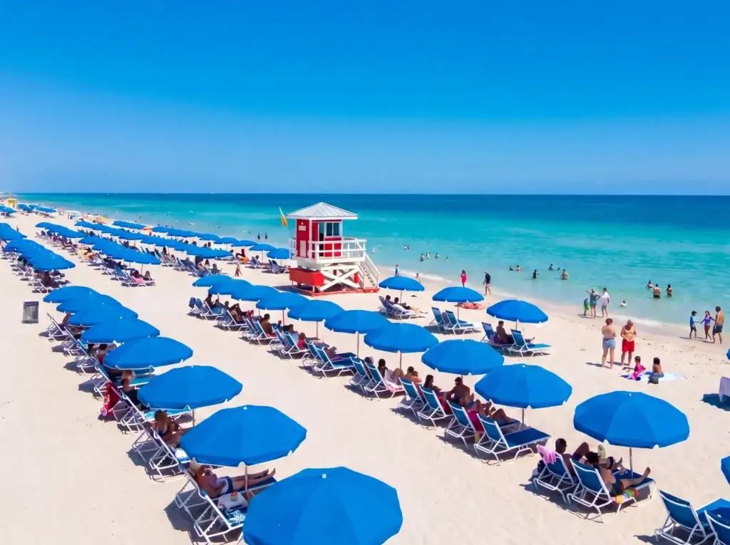 Busy Fort Lauderdale Beach at midday with rows of blue beach umbrellas and lounge chairs, a red lifeguard stand, and beachgoers wading in the turquoise Atlantic Ocean under a clear blue sky